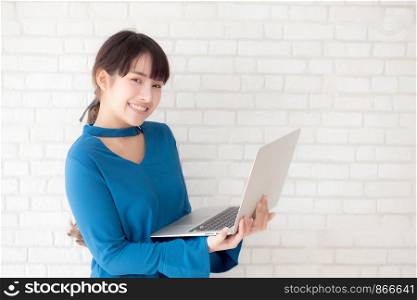 Beautiful portrait asian young woman smile using laptop standing at workplace on cement concrete background, girl happy with computer internet online, lifestyle and freelance business concept.