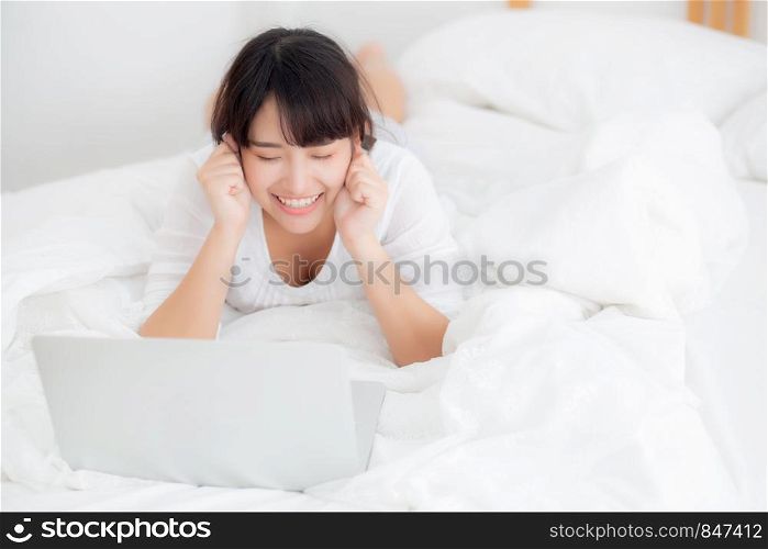 Beautiful portrait freelance asian young woman lying relax and leisure working laptop computer internet online on bed in bedroom, cheerful asia girl with gesture glad and success, lifestyle concept.