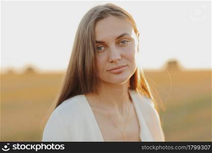 Beautiful portrait of a young woman during the sunset with warm yellow sun rays on her face with bales of straw on the background.. Beautiful portrait of a young woman during the sunset with warm yellow sun rays on her face with bales of straw on the background