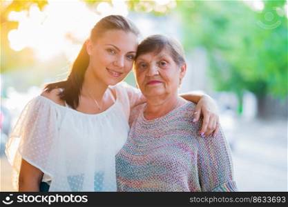 Beautiful portrait of grandmother and her granddaughter standing outdoor on summer street. Golden hour, sun flares. Girl embracing granny with love. Family concept.. Beautiful portrait of grandmother and her granddaughter standing outdoor on summer street. Golden hour, sun flares. Girl embracing granny with love. Family concept