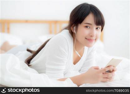 Beautiful portrait young asian woman lying happy earphone listening music online with relax and enjoy in the bedroom, girl fun headphone with playing song smart mobile phone, lifestyle leisure concept.