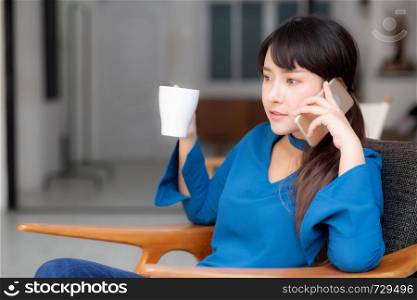 Beautiful portrait young asian woman smiling using mobile smart phone talking enjoy and drink coffee with relax, girl sitting on chair at living room, communication concept.