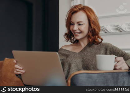 Beautiful positive redhead woman happy to have video call with best friend, share news with each other, drinks coffee, sits in comfortable armchair, watches intereting movie, connected to internet