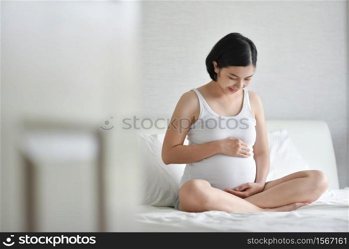 Beautiful pregnant Asian woman in white Pajamas smiling sitting on bed. Healthy Happy Young mother holding and touching and looking her belly at home while pregnancy. Family Maternity lifestyle.