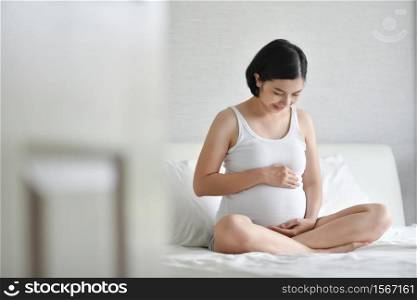 Beautiful pregnant Asian woman in white Pajamas smiling sitting on bed. Healthy Happy Young mother holding and touching and looking her belly at home while pregnancy. Family Maternity lifestyle.