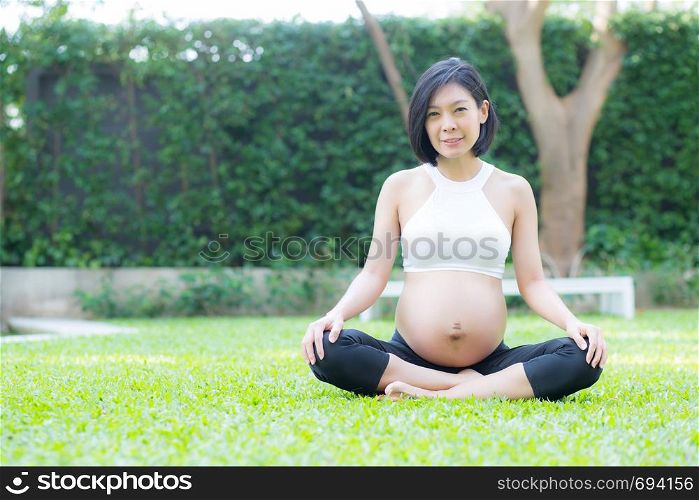 Beautiful pregnant asian young woman relax in the park, girl with belly sitting with crossed leg on grass and yoga exercise for wellness, copy space.