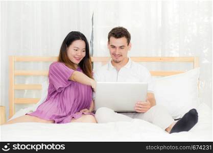 Beautiful pregnant family with laptop computer on bedroom, husband and wife notebook relax together, couple and family concept.