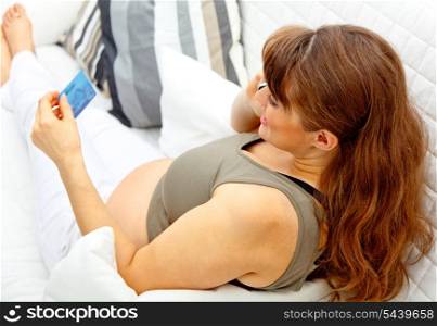 Beautiful pregnant female with mobile phone and credit card on sofa at home&#xA;
