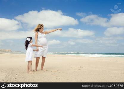 Beautiful pregnant woman and her little daughter on the beach pointing to the sea