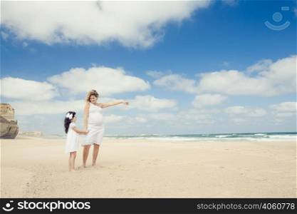 Beautiful pregnant woman and her little daughter on the beach pointing to the sea
