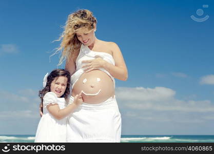 Beautiful pregnant woman in the beach with her little daugther making a smile on mom&rsquo;s belly with sunscreen