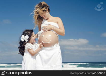 Beautiful pregnant woman in the beach with her little daugther making a smile on mom&rsquo;s belly with sunscreen