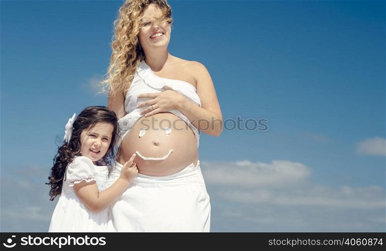 Beautiful pregnant woman in the beach with her little daugther making a smile on mom&rsquo;s belly with sunscreen
