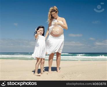 Beautiful pregnant woman in the beach with her little daugther making a smile on mom&rsquo;s belly with sunscreen