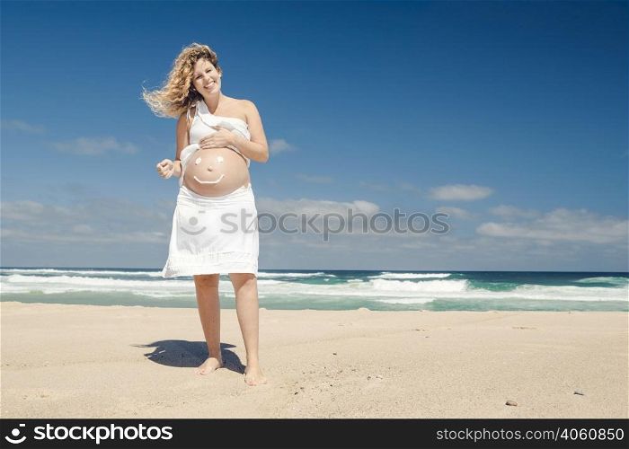Beautiful pregnant woman in the beach with smile on the belly made with sunscreen