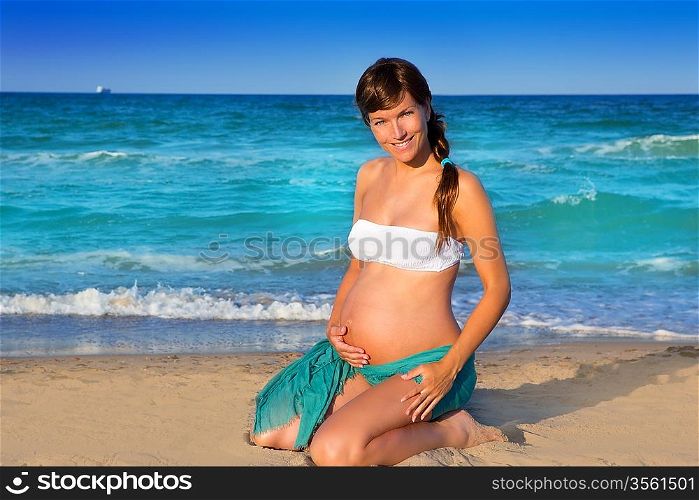 Beautiful pregnant woman on knees at blue beach sand in summer vacation