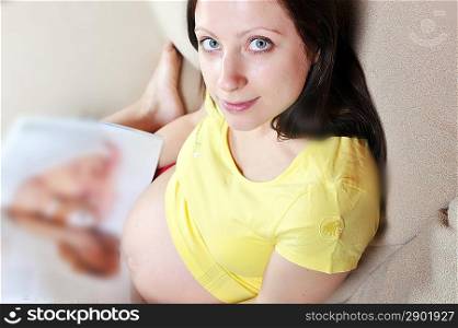 Beautiful pregnant woman relaxing on couch and reading magazine