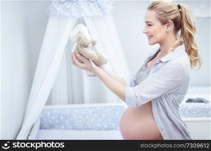 Beautiful pregnant woman standing in the child’s room with soft toys in hands, decorates and fixing everything in the bedroom for her infant, care of baby comfort  