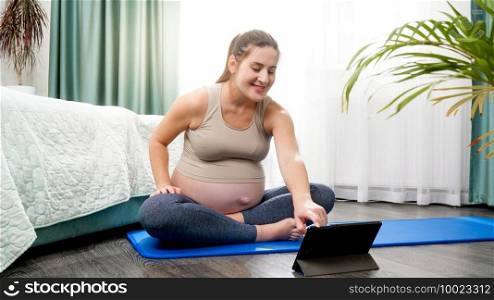 Beautiful pregnant woman stretching and doing fitness exercises on online yoga lesson via tablet computer. Concept of healthy lifestyle, healthcare and sports during pregnancy.. Beautiful pregnant woman stretching and doing fitness exercises on online yoga lesson via tablet computer. Concept of healthy lifestyle, healthcare and sports during pregnancy