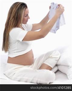 Beautiful Pregnant Young Woman Holding Up New Baby Clothes, Waiting For Her Baby To Be Born