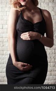 Beautiful pregnant young woman in a black tight dress against a white brick wall