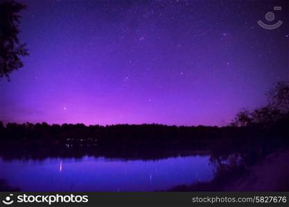 Beautiful purple night sky with many stars on a lake with forest on the other coast. Milkyway reflection in water