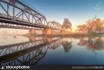Beautiful railroad bridge and river in fog at sunrise in autumn. Industrial landscape with railway station, reflection in water, road, mist, trees, blue sky in foggy morning in fall. Transportation. Beautiful railroad bridge and river in fog at sunrise in autumn