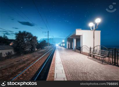 Beautiful railway station at night in summer. Rural railroad at dusk. Industrial landscape with railroad, railway platform, city lamps, buildings, trees, blue sky with clouds at twilight. Travel. Railway station at night in summer. Starry sky over railroad at dusk