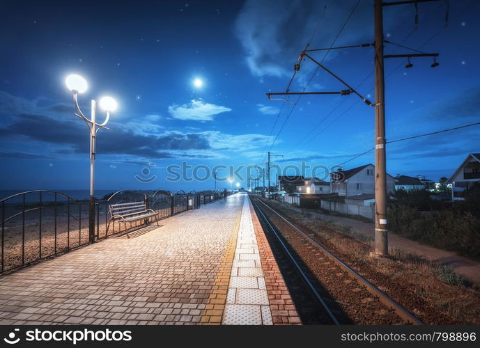 Beautiful railway station at night in summer. Starry sky over railroad at dusk. Industrial landscape with railroad, railway platform, city lights, buildings, blue sky with moon and clouds at twilight. Railway station at night in summer. Starry sky over railroad