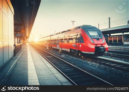 Beautiful railway station with modern high speed red commuter train at colorful sunset. Railroad with vintage toning. Train at railway platform. Industrial concept. Railway tourism
