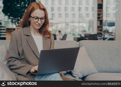 Beautiful red-haired business woman in casual clothes and eyeglasses working online, using laptop, sitting on comfortable grey couch in office lounge, female entrepreneur enjoying distant work. Woman working remotely in coworking space, using laptop while sitting on couch