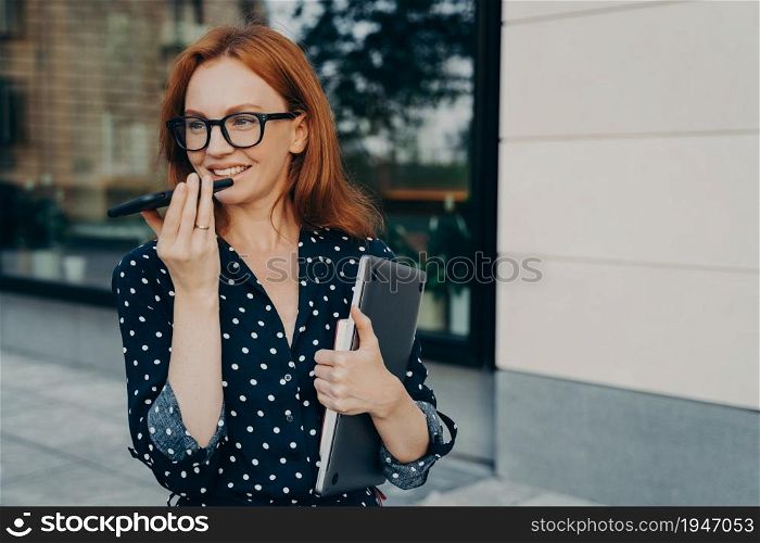 Beautiful red-haired business woman stands outdoors with mobile phone and records audio message or smartphone, talks with virtual digital voice assistant on way to office, female using speakerphone. Beautiful business woman stands outdoors with mobile phone and records audio message or smartphone