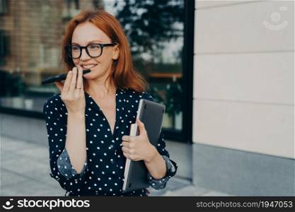 Beautiful red-haired business woman stands outdoors with mobile phone and records audio message or smartphone, talks with virtual digital voice assistant on way to office, female using speakerphone. Beautiful business woman stands outdoors with mobile phone and records audio message or smartphone