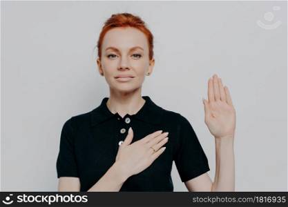 Beautiful red-haired patriotic woman wearing black t-shirt swearing with hand on chest and open palm, making loyalty promise oath while posing isolated over grey studio background. Beautiful red haired patriotic woman swearing with hand on chest and open palm