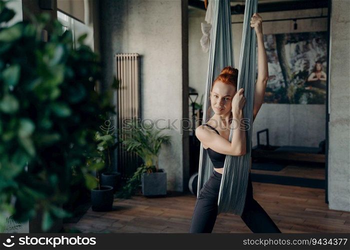 Beautiful red-haired woman in active wear rests after fly yoga in fitness studio. Relaxing on a grey hammock, smiling at the camera. Embracing health, sport, and yoga.