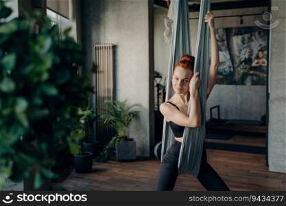 Beautiful red-haired woman in active wear rests after fly yoga in fitness studio. Relaxing on a grey hammock, smiling at the camera. Embracing health, sport, and yoga.