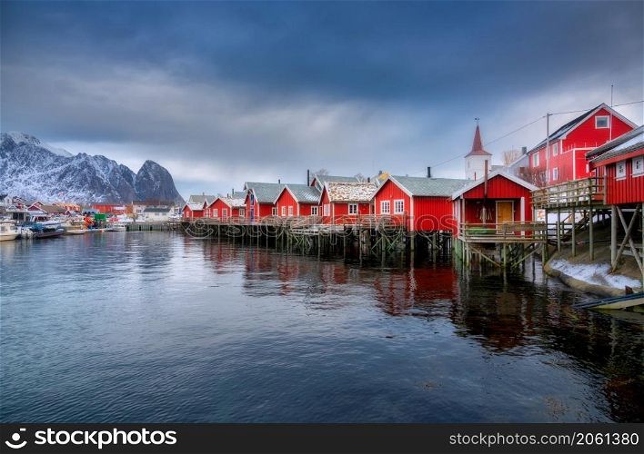 Beautiful red rorbu in overcast day. Fishing village in Lofoten islands, Norway. Winter landscape with houses, snowy mountains, sea, sky with clouds. Norwegian traditional red rorbuer on the water