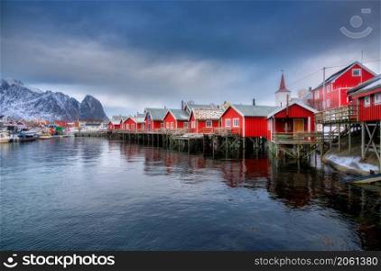 Beautiful red rorbu in overcast day. Fishing village in Lofoten islands, Norway. Winter landscape with houses, snowy mountains, sea, sky with clouds. Norwegian traditional red rorbuer on the water