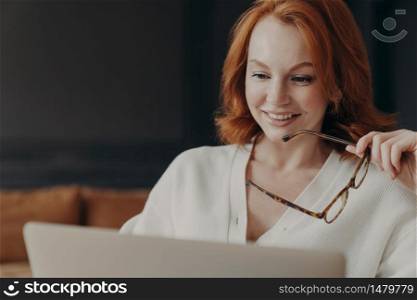 Beautiful redhaired woman concentrated in laptop display, smiles positively, holds optical glasses, poses indoor, uses modern technologies for online communication, watches webinar, reads news