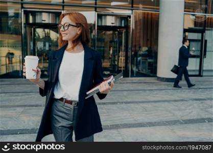 Beautiful redhead female entrepreneur standing outdoors with coffee cup and laptop in hands, leaving office, businesswoman in spectacles dressed in formal clothes waiting for taxi and looking aside. Beautiful redhead female entrepreneur standing outdoors with coffee cup and laptop in hands