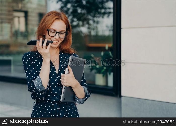 Beautiful redhead female entrepreneur walks outdoor records voice message in social media via smartphone carries laptop uses modern gadgets for communication poses near city building. Lifestyle. Female entrepreneur walks outdoor records voice message in social media via smartphone