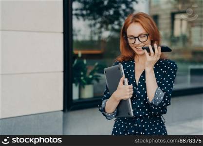 Beautiful redhead female entrepreneur walks outdoor records voice message in social media via smartphone carries laptop uses modern gadgets for communication poses near city building. Lifestyle. Female entrepreneur walks outdoor records voice message in social media via smartphone