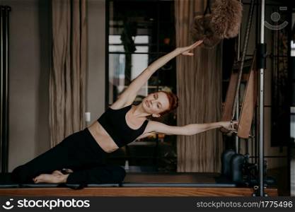 Beautiful redhead woman pilates trainer exercising and stretching out on pilates cadillac with arms outstreched to one side, showing flexibility of perfect body. Fitness, healthy lifestyle and sport. Beautiful woman exercising and stretching on cadillac reformer