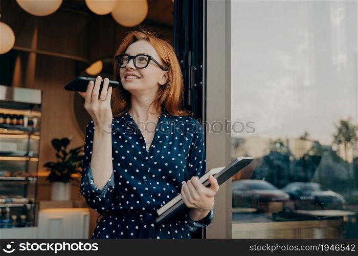 Beautiful redhead woman speaks on speaker via smartphone records voice message looks away uses translationn app wears fashionable formal clothes. Voice recognition and distant communication.. Pretty woman speaks on speaker via smartphone records voice message