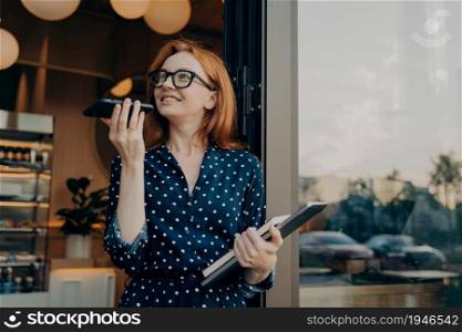 Beautiful redhead woman speaks on speaker via smartphone records voice message looks away uses translationn app wears fashionable formal clothes. Voice recognition and distant communication.. Pretty woman speaks on speaker via smartphone records voice message