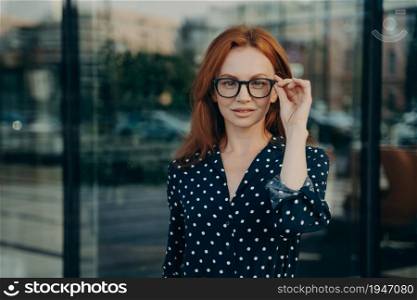 Beautiful redhead woman wears transparent glasses and polka dot dress looks directly at camera poses near office building against blurred background. Businsswoman in formal wear stands outdoor. Beautiful redhead woman poses near office building against blurred background