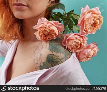 Beautiful redhead woman with living coral colored flowers on her shoulders with tattoo roses on a blue background. Greeting holiday card for Mother's or Woman's Day.. Young cute girl with tattoo on her shoulders and a bouquet of coral roses on a blue background. Postcard to Mother's day.