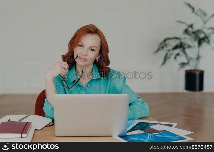 Beautiful redhead woman works distantly from home, sits in front of laptop computer finishes working on project thinks about adding interesting content on own website prepares publication for web page