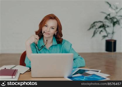 Beautiful redhead woman works distantly from home, sits in front of laptop computer finishes working on project thinks about adding interesting content on own website prepares publication for web page