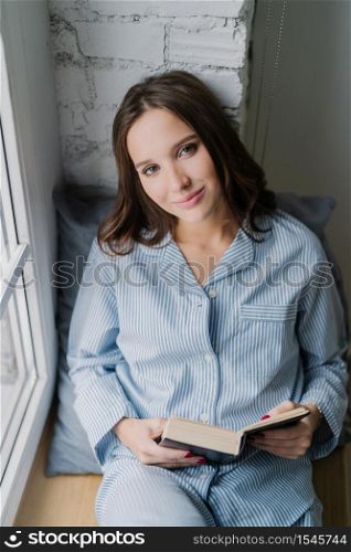 Beautiful relaxed woman with carefree expression, dressed in nightclothes, reads book on window sill in morning, enjoys spare time, likes her hobby. Vertical shot of teenage girl with literature